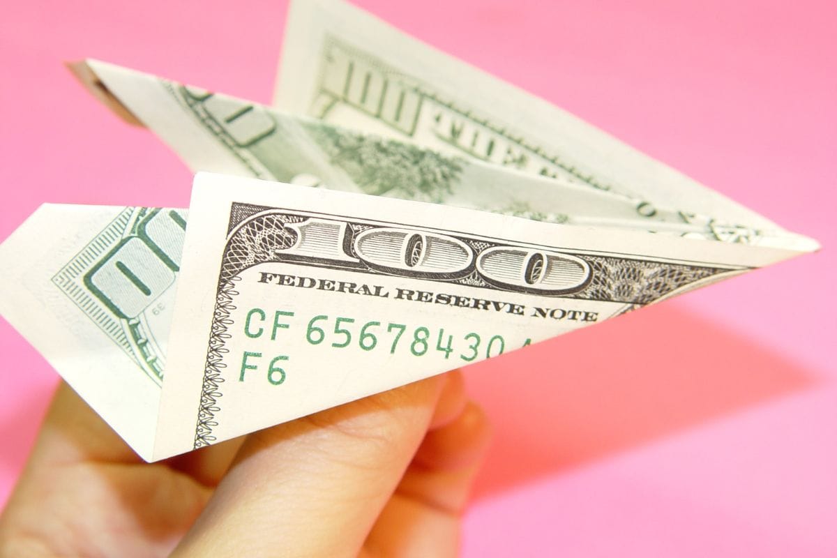  A hand holding an origami airplane made of a 100 dollar bill against a pink background, symbolizing the benefits of saving money for future.